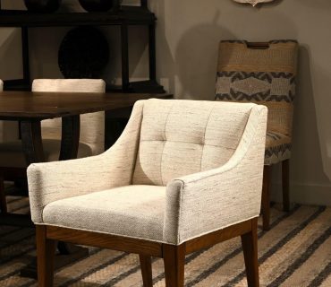 a cozy living space with a beige upholstered armchair, a geometric patterned rug, shelving with decor items, and a circular wall mirror